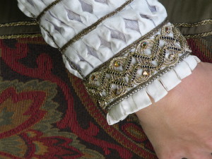 The puffs were mounted to the base layer and sewn down along the horizontal lines. Then the edges of each strip were caught back in the middle. Gold trim was sewn over the horizontal stitching lines, and small, looped pickadils of white silk sewn to the wrists of the sleeves. All sewing was done by hand with gold-colored silk thread.